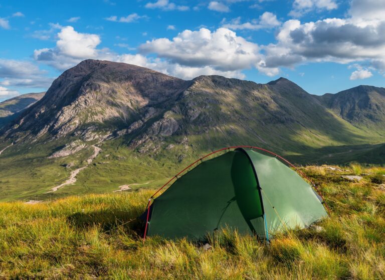 Green dome tent pitched on a grassy hillside overlooking sunlit rugged mountains beneath a blue sky dotted with white clouds.