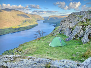 Green backpacking tent pitched on a grassy cliff ledge beside rocky outcrops, overlooking a long blue lake winding between sunlit hills and a lone bare tree under a partly cloudy sky.