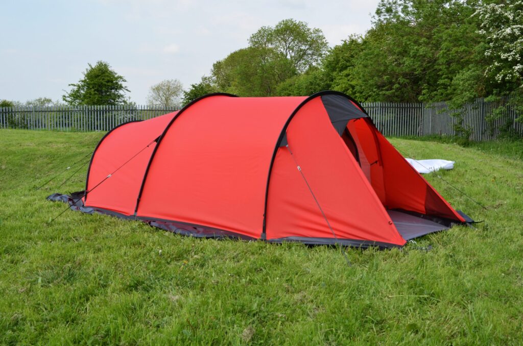 Red tunnel-style camping tent with black trim and guy lines staked on a grassy field, open entrance to the right and trees and a metal fence in the background