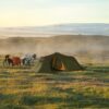 Green tunnel-style tent pitched on a dewy grassy plain at sunrise, two loaded touring bicycles with panniers parked beside it amid low mist and rolling hills with a distant snow-covered ridge.