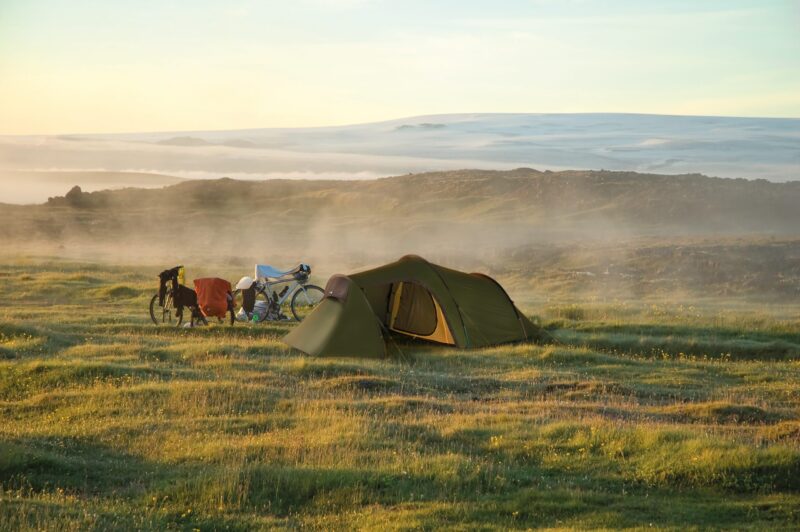 Green tunnel-style tent pitched on a dewy grassy plain at sunrise, two loaded touring bicycles with panniers parked beside it amid low mist and rolling hills with a distant snow-covered ridge.