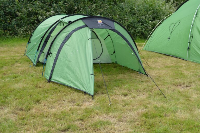 Green tunnel-style camping tent with its entrance open, held by flexible poles and guy ropes on a grassy field with shrubs behind and a second green tent partly visible at the right