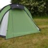Large light-green dome camping tent with a small black canopy and black trim staked on a grassy field, with a hedgerow of white-flowering bushes behind and a smaller green tent partly visible at the right edge.