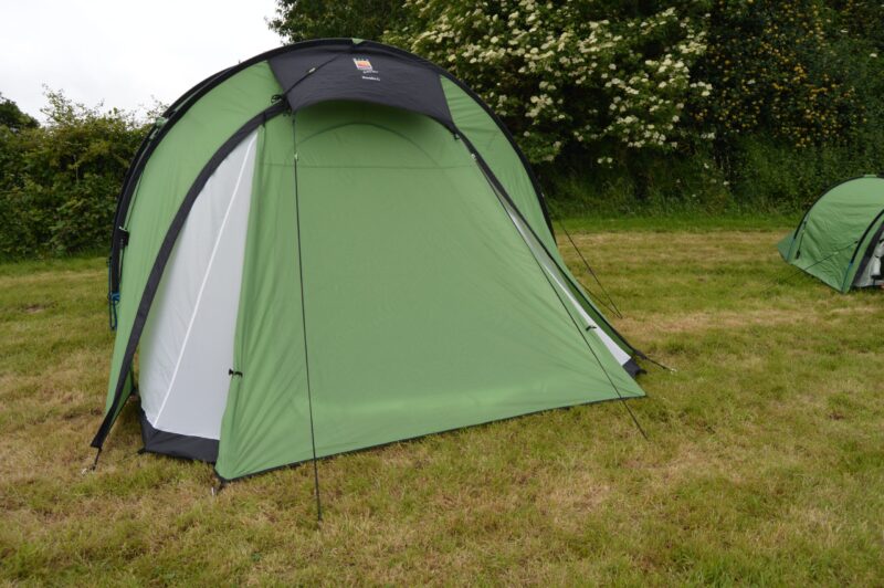 Large light-green dome camping tent with a small black canopy and black trim staked on a grassy field, with a hedgerow of white-flowering bushes behind and a smaller green tent partly visible at the right edge.