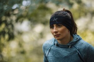 Young woman in a blue athletic hoodie and a black headband with a logo, hair tied back in a ponytail, looking thoughtfully to the side against a softly blurred green outdoor background.