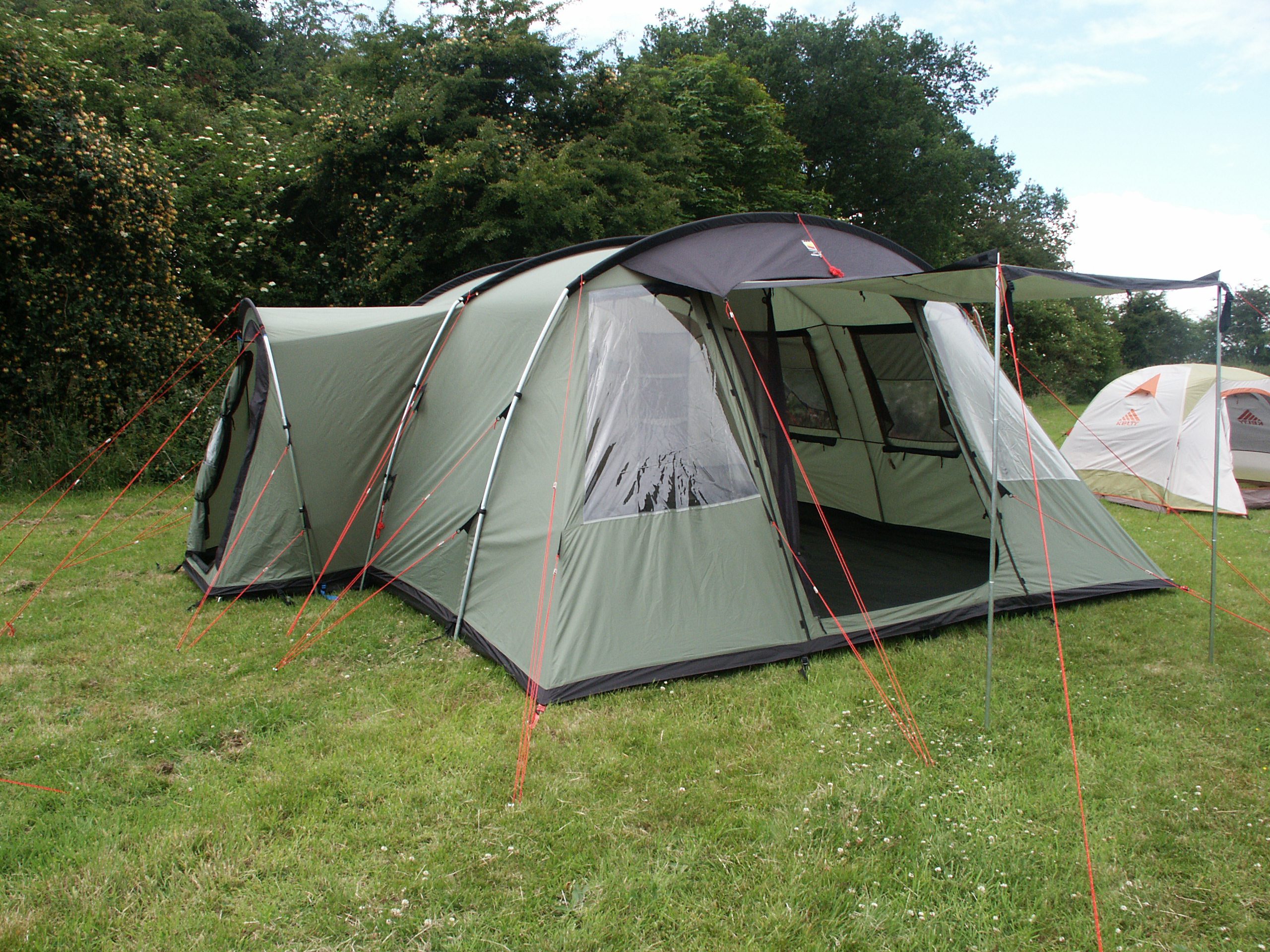 Large green multi-room family tent with an open front awning and clear side window, held by red guy lines on a grassy campsite with trees behind and a smaller white tent to the right.