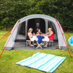 Adults and children sit around a small table inside a grey dome tent with red guy lines, sharing a meal while a striped blue-and-white picnic blanket lies on the grass in front and two blue folding camping chairs sit to the right against a hedge backdrop.