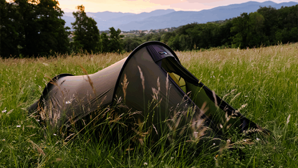 Starlite-1 Olive-green tunnel tent pitched in tall meadow grass at dusk, with trees nearby and rolling blue mountains on the horizon.
