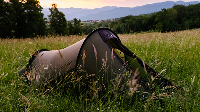 Olive-green tunnel tent pitched in tall meadow grass at dusk, with trees nearby and rolling blue mountains on the horizon.