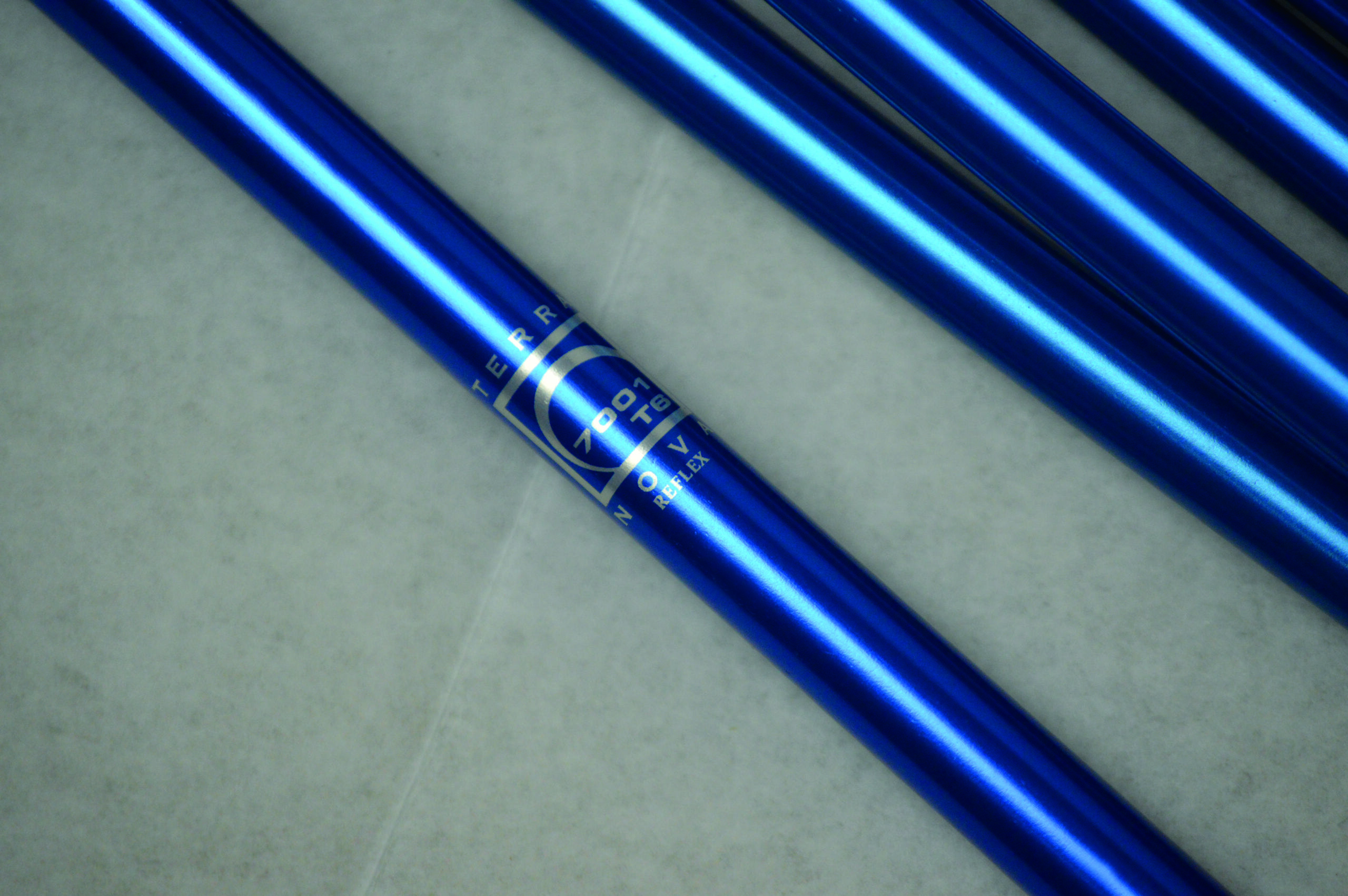 Diagonal close-up of several shiny metallic blue aluminium tubes on a pale textured surface, one tube showing a white circular logo and small printed text.