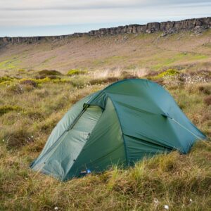 Green two‑person tent pitched in windswept grassy moorland dotted with white cotton‑grass, with a low rocky escarpment and pale cloudy sky in the background.