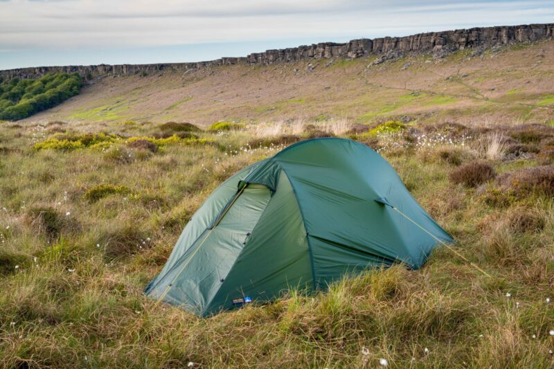 Terra Nova Shoot Green two‑person tent pitched in windswept grassy moorland dotted with white cotton‑grass, with a low rocky escarpment and pale cloudy sky in the background.