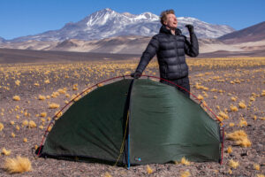 A camper in a black puffer jacket and gloves stands laughing with a raised hand behind a small green dome tent on a rocky high-altitude plain dotted with yellow tussock grass, snow-capped mountains and a clear blue sky in the background.