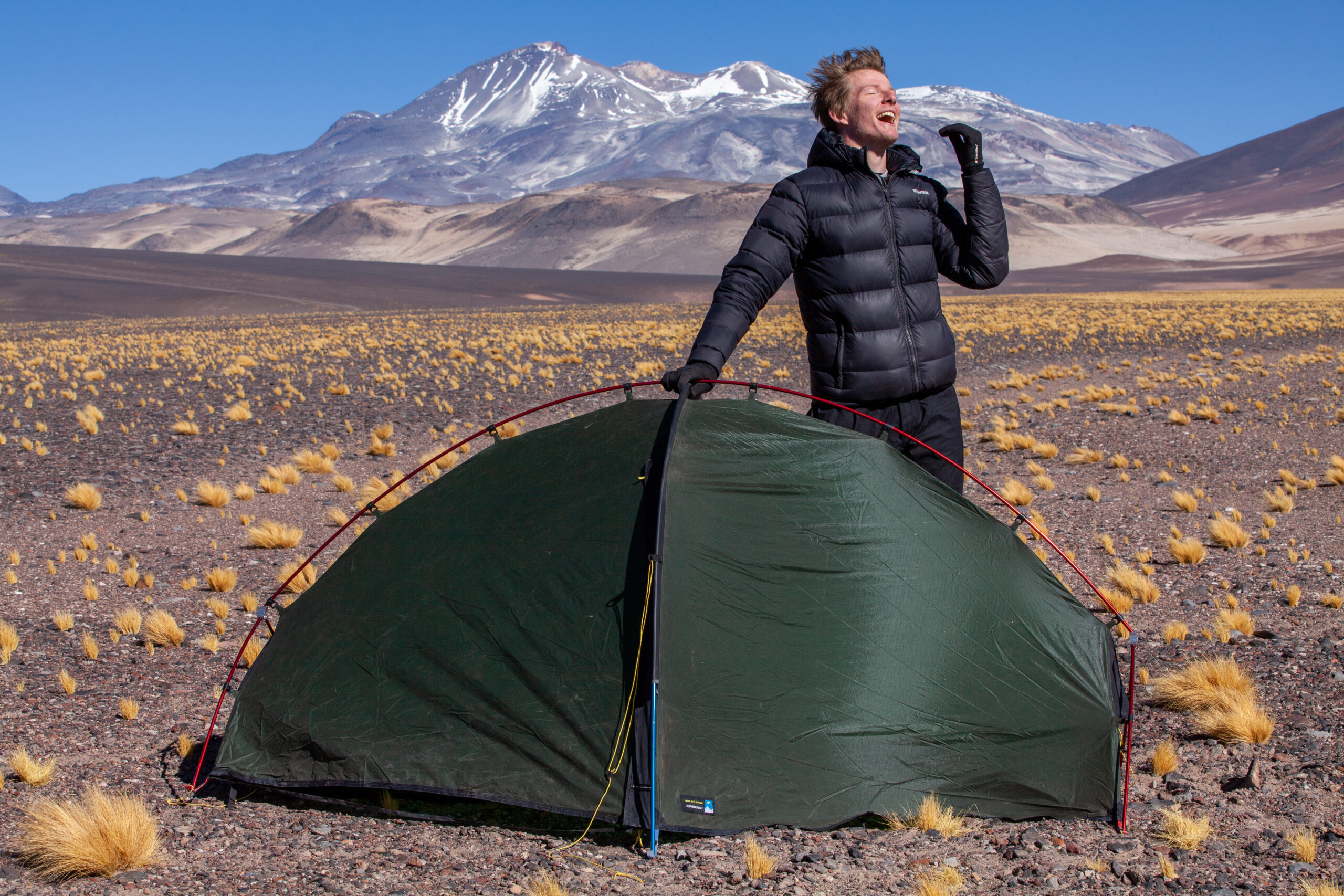 A camper in a black puffer jacket and gloves stands laughing with a raised hand behind a small green dome tent on a rocky high-altitude plain dotted with yellow tussock grass, snow-capped mountains and a clear blue sky in the background.
