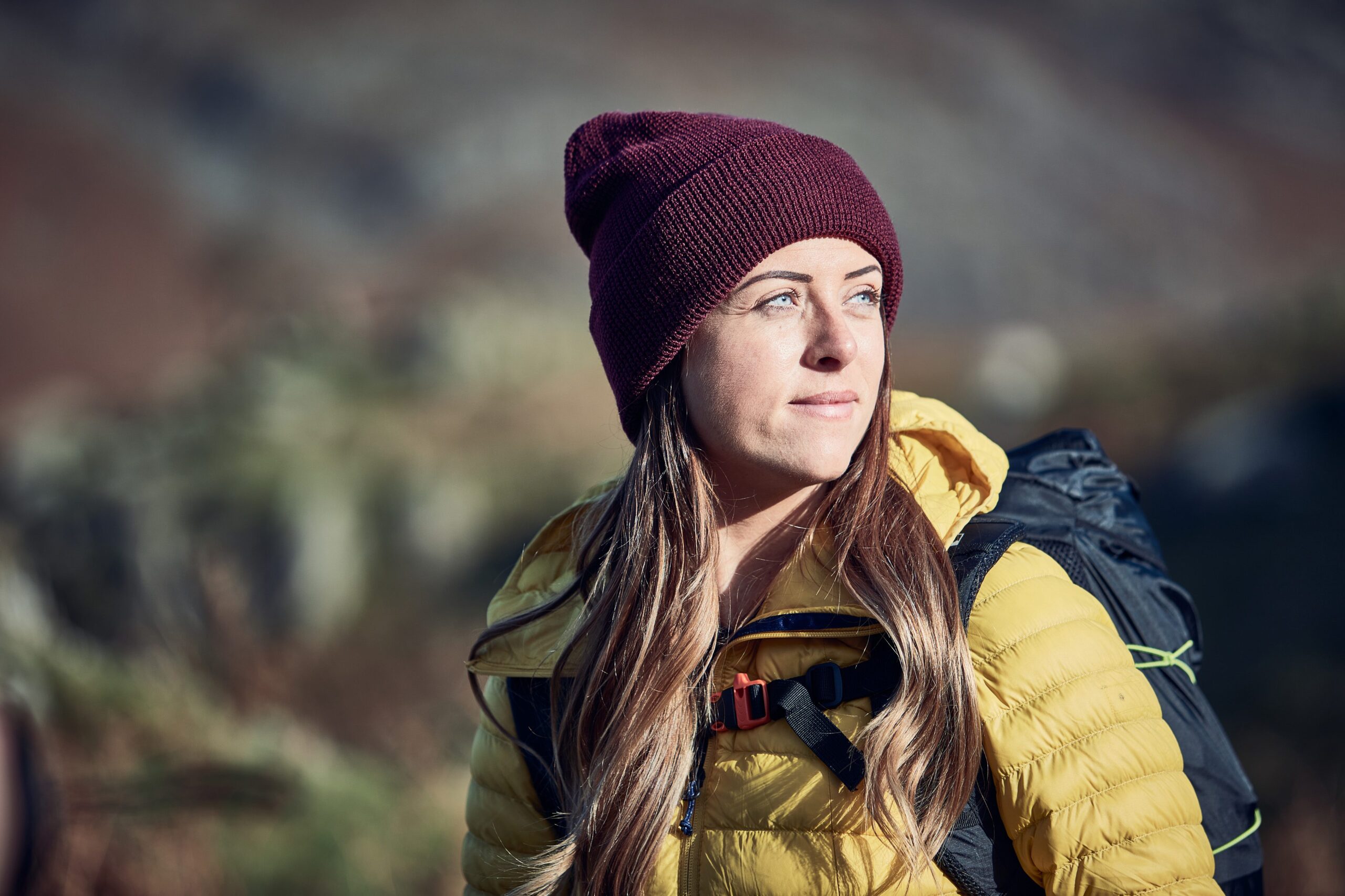 Young woman wearing a maroon knit beanie and yellow puffer jacket with a backpack looks to the right as soft sunlight illuminates her face against a blurred mountainous outdoor background.