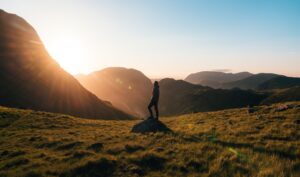 Silhouette of a person standing on a rock, overlooking a breathtaking mountain landscape at sunset, with warm sunlight illuminating the scene and highlighting the rolling hills and valleys.