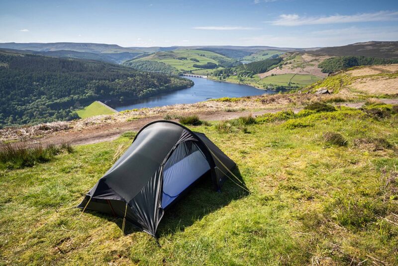Laser Compact AS (2) Small dark backpacking tent pitched on a grassy hilltop overlooking a winding blue reservoir and green rolling hills under a clear sky.