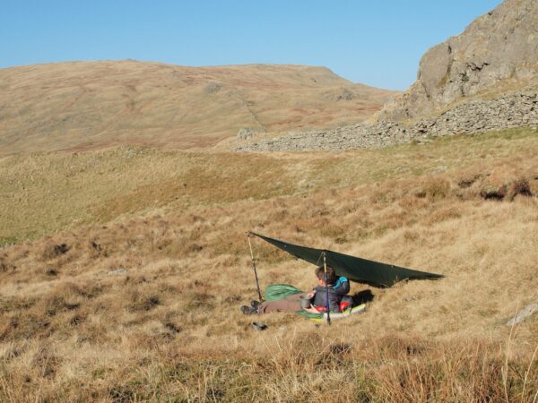 TARP 1 A person sitting under a makeshift shelter in a grassy, hilly landscape on a clear day, surrounded by rolling hills and rocky outcrops.