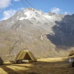 Two khaki tents pitched on a grassy alpine plateau beneath steep, snow-dusted jagged peaks with cable lines overhead, and a person in a red jacket sitting on the right.