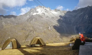 Two khaki tents pitched on a grassy alpine plateau beneath steep, snow-dusted jagged peaks with cable lines overhead, and a person in a red jacket sitting on the right.