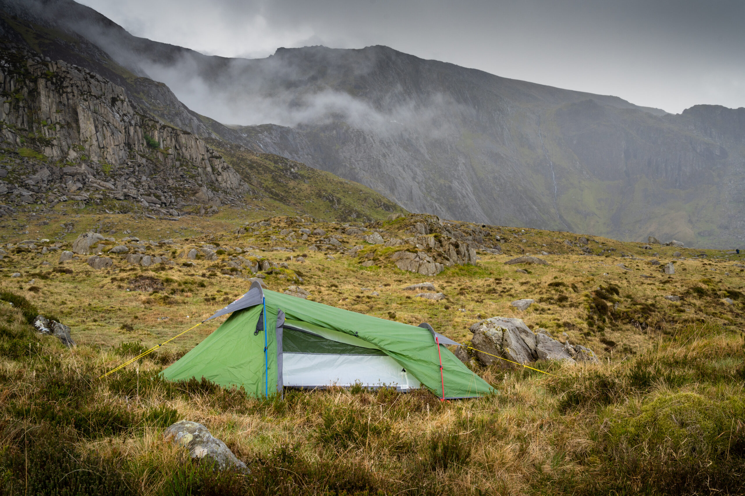 Green camping tent set up in a rugged mountain landscape with misty peaks in the background, showcasing a tranquil outdoor adventure setting.