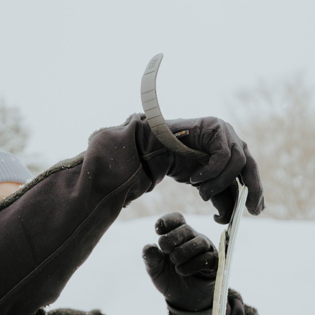 Gloved hands in black winter gloves adjusting a snowboard binding strap on the edge of a snowboard in a snowy outdoor setting with blurred trees in the background.
