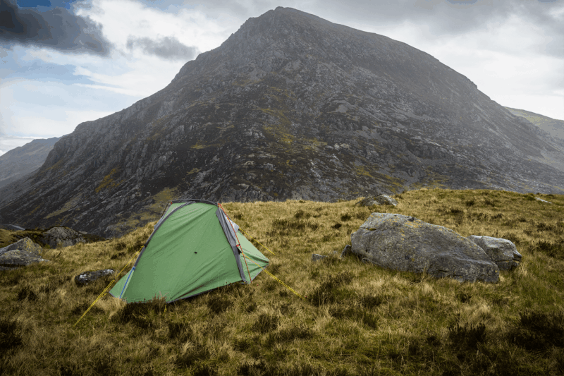 Helm-Compact-1-4_LR A small green tent with yellow guy lines pitched on grassy highland beside large grey boulders, with a steep rocky mountain filling the background under a cloudy sky.