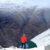 Southern-Cross-1 Person in an orange jacket and knit hat stands beside a green tent on a snowy mountainside, looking down into a steep, snow-dusted valley with rugged brown rock slopes.