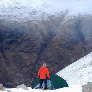 Person in an orange jacket and knit hat stands beside a green tent on a snowy mountainside, looking down into a steep, snow-dusted valley with rugged brown rock slopes.