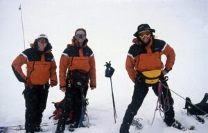 Three mountaineers in bright orange jackets stand on a snowy peak, equipped with climbing gear and ice axes, showcasing their adventurous spirit in extreme weather conditions.
