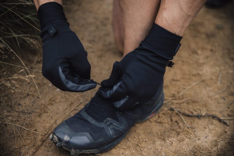 Hands in black gloves tightening a quick‑pull lace on a worn black trail running shoe on sandy ground beside dry grass.