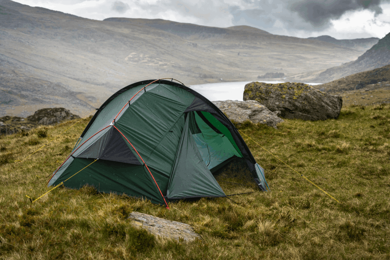Southern-Cross-2-2_LR Green dome tent with red poles and yellow guy lines pitched on a grassy hillside beside a lake, with large boulders and misty rolling hills under a cloudy sky