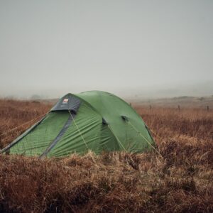 Green dome tent pitched in windswept brown moorland with yellow guy lines, surrounded by tall dry grass under a foggy, overcast sky.
