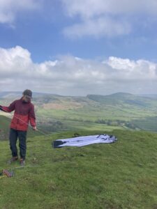 Person in a red jacket and cap holding tent poles beside a grey tent body spread on a grassy hilltop, overlooking rolling green valleys under a partly cloudy blue sky.