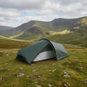 A green camping tent set up on a grassy field surrounded by rolling hills and mountains under a partly cloudy sky, ideal for outdoor adventures and hiking trips.