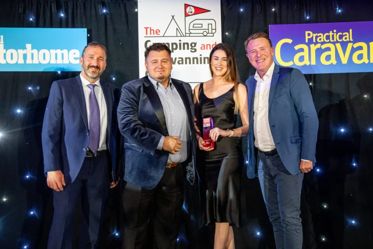Four formally dressed people smiling on a stage in front of Camping and Caravanning and Practical Caravan signs, with a woman in a black dress holding a red award trophy.