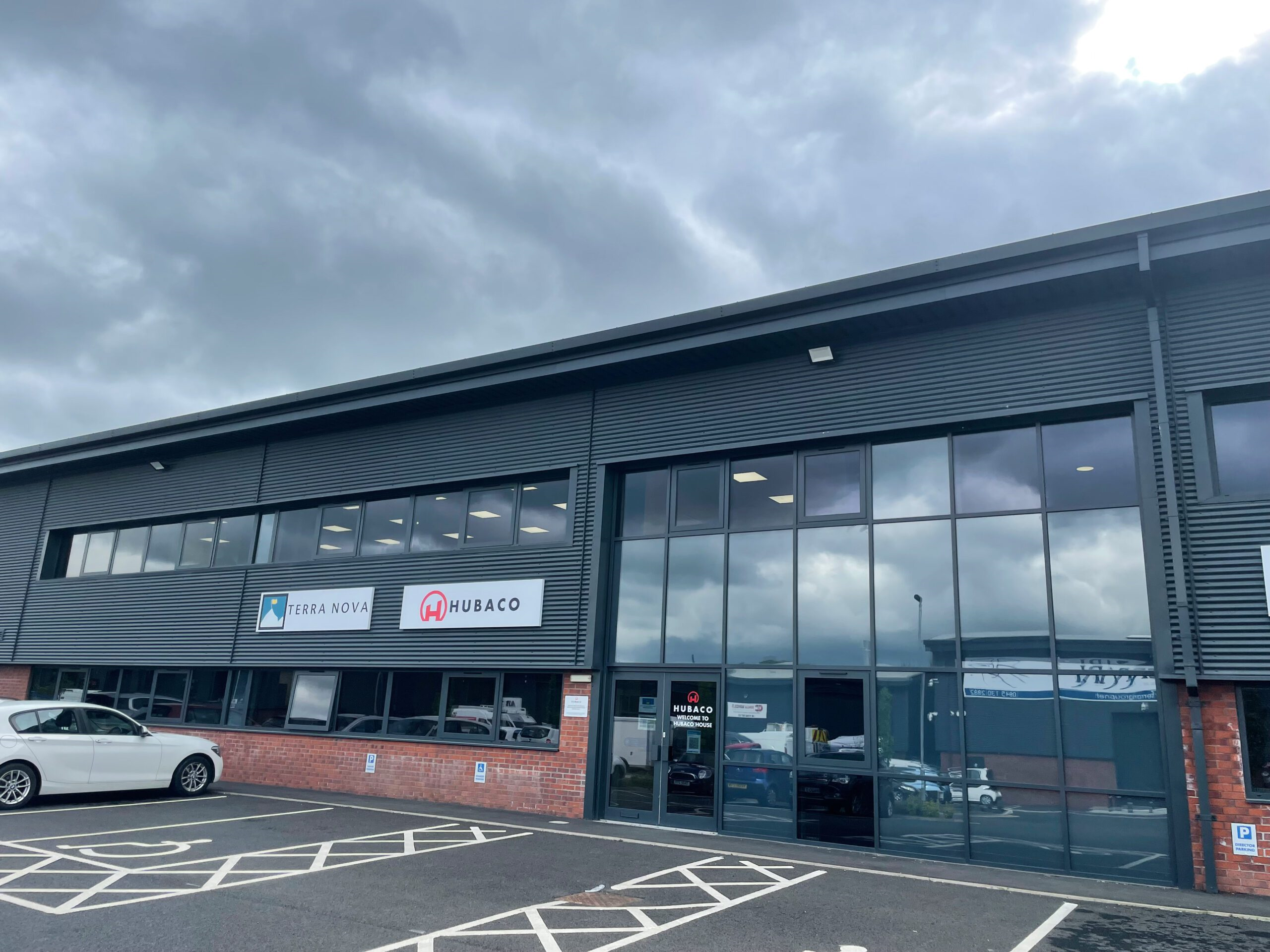 Low-rise dark-grey industrial office building with large floor-to-ceiling windows and signs reading Terra Nova and Hubaco above a brick base, a white car parked by marked bays and a cloudy sky overhead.