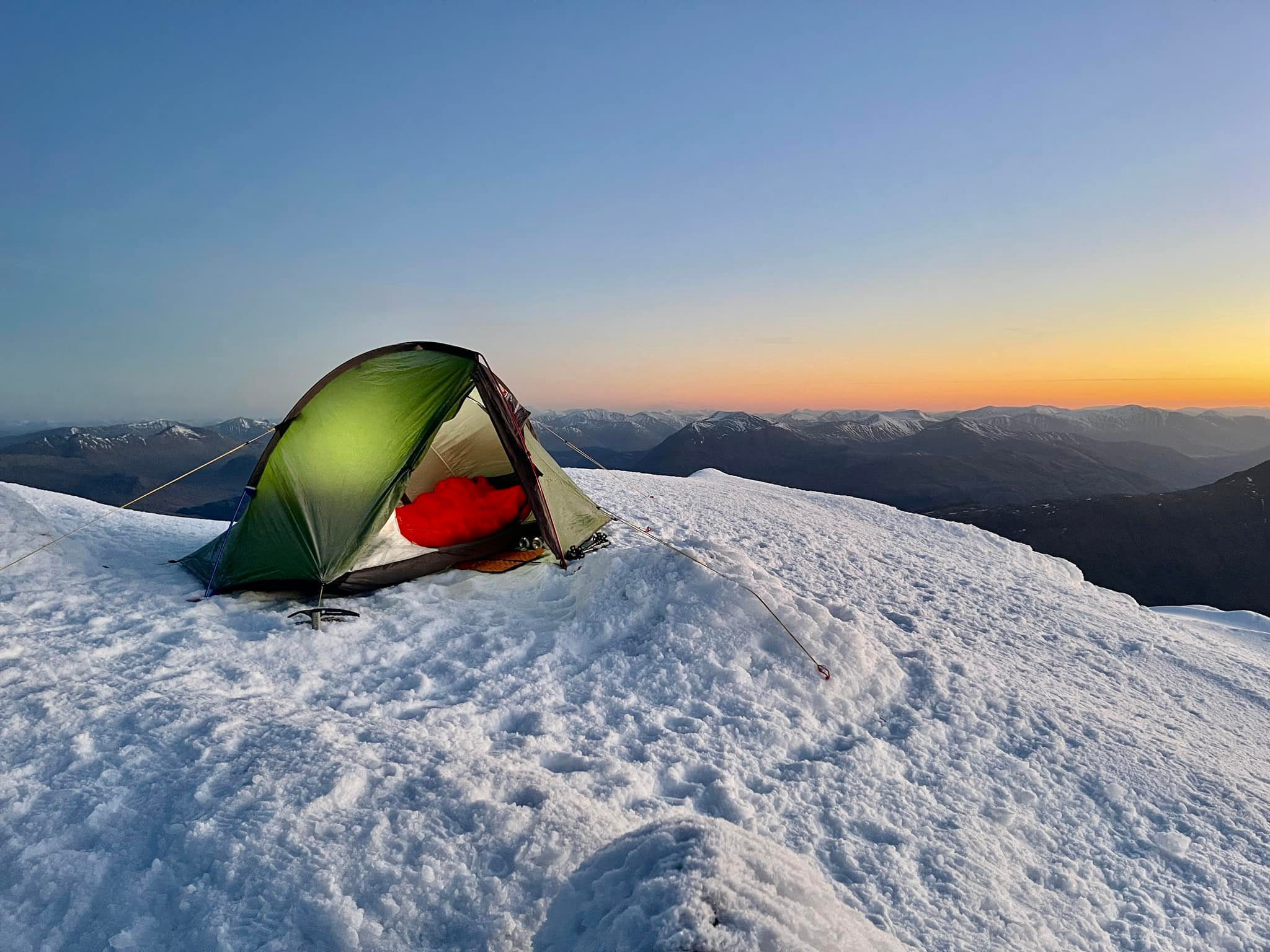 Green two-person tent pitched on a snowy mountain summit at sunrise, a red sleeping bag visible inside, guy lines and an ice axe anchoring it, with distant snow-capped peaks under a clear orange-blue sky.