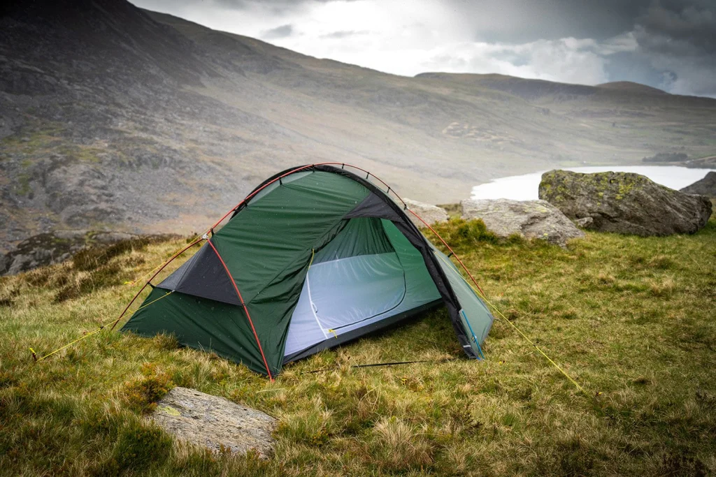 Green dome tent with red poles and an open inner mesh pitched on a windswept grassy hillside among rocks, overlooking a small lake and misty hills under an overcast sky.