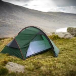 Green dome tent with red poles and an open inner mesh pitched on a windswept grassy hillside among rocks, overlooking a small lake and misty hills under an overcast sky.
