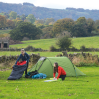 Two people setting up camp beside a green tent in a grassy field—one kneeling in a red jumper arranging a sleeping mat and the other in a navy hoodie holding a black-and-red sleeping bag—with a dry-stone wall and tree-covered rolling hills in autumn colours behind them.
