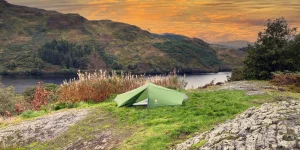 Green two-person tent pitched on a grassy rocky ridge beside tall dried wildflowers, overlooking a calm lake and tree-covered hills beneath an orange sunset sky.