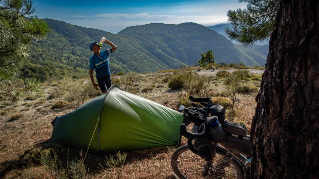 Cyclist in a blue jersey drinks from a water bottle beside a small green tent and a loaded touring bike on a sunlit mountain ridge with forested hills and pine trees beyond.