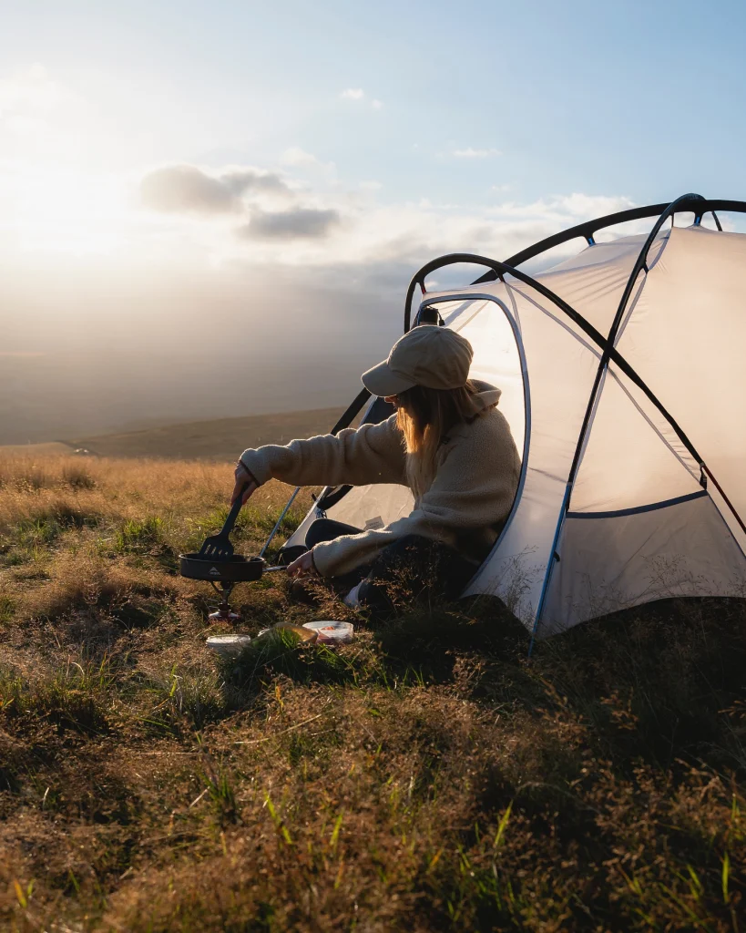 Person in a cap and fleece sitting at the entrance of a light-coloured tent on a grassy hillside, stirring a small frying pan on a portable camping stove as golden sunlight falls over rolling hills.