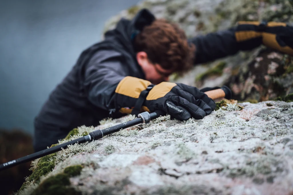 A climber's gloved hand (black and yellow, 'extremities' logo visible) grips a frost-dusted rocky ledge beside a trekking pole, the climber blurred in the background.