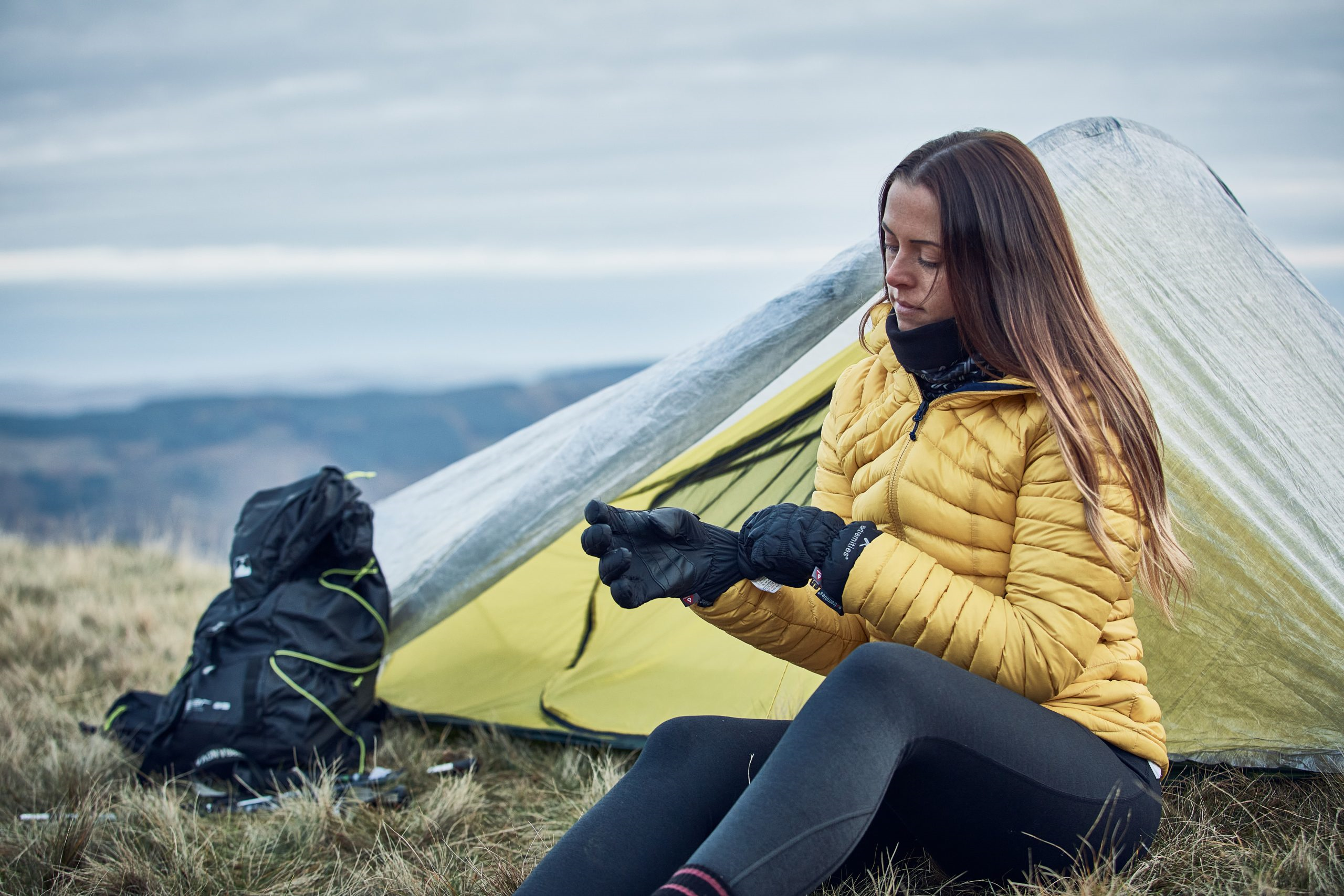A woman in a yellow insulated jacket sits outside a lightweight tent on a grassy hillside, pulling on black gloves beside a backpack with distant rolling hills under a cloudy sky.