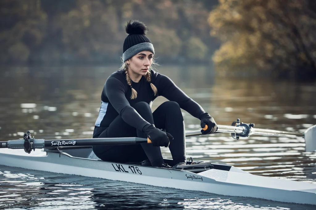 Woman in a black thermal outfit and grey-and-black pom-pom beanie with braided hair, pulling oars on a single scull across a calm lake with blurred autumn trees behind her