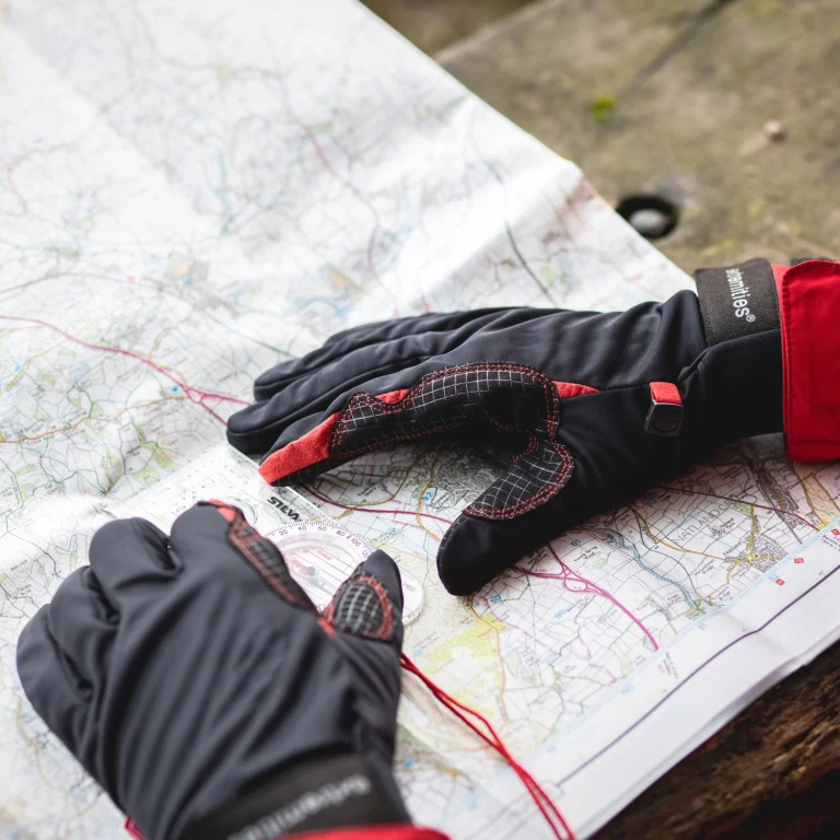 ls_ex_os_2021 Pair of black and red outdoor gloves resting on an unfolded topographic map, one glove showing textured palm stitching and the other near a clear baseplate compass with a red lanyard on a wooden bench.