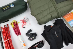 Flat-lay of camping gear on a white background showing a green Terra Nova Laser Compact 1 sleeping bag in its stuff sack, an olive quilted jacket, black gloves, sunglasses, a headlamp, a clear-base compass with red cord, bundles of red tent poles and pegs, a bottle of SPF50 sunscreen and folded maps.