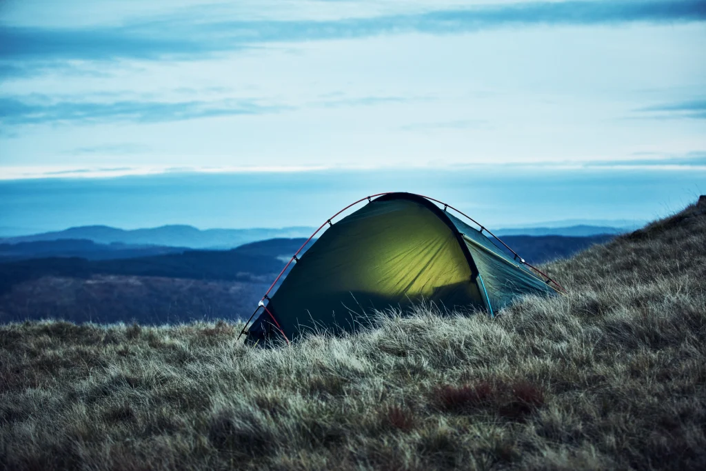 A green dome tent lit from within, pitched on a windswept grassy hillside at dusk with layered blue hills and a wide sky in the background.